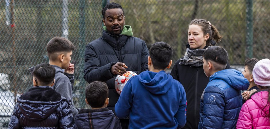 ein Sporttrainer mit einem Ball in der Hand und eine Gesundheitsberaterin sprechen mit einer Gruppe von Kindern auf der Straße. 