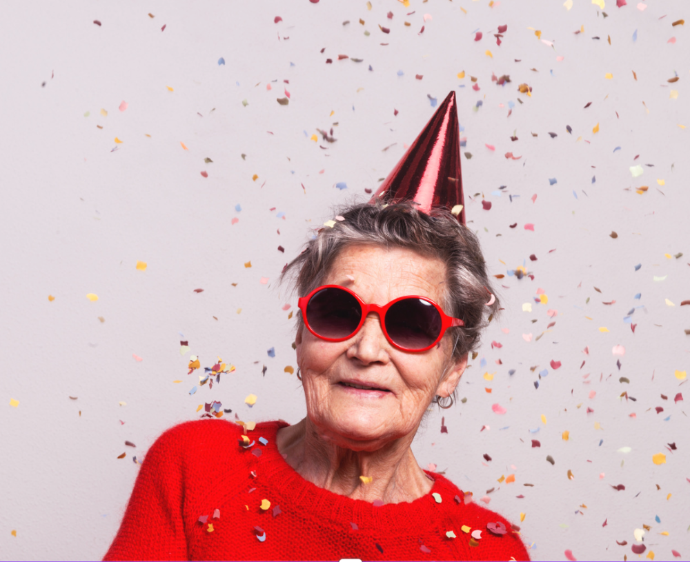 Das Bild zeigt eine ältere Frau mit einem typischen Festhut (spitzer, farbiger Papierhut), Sonnenbrille und Konfetti im Hintergrund.